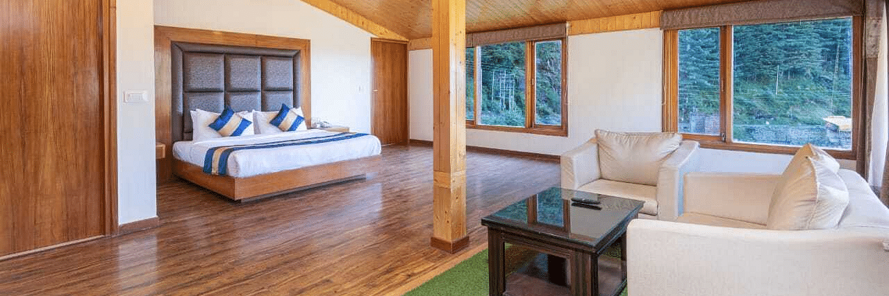 The image shows a spacious hotel suite with a king size bed, a sitting area, and large windows with mountain views  | DLS Serenity Resort and Spa